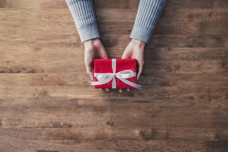 Woman hands in gray sweater on wood table giving red Christmas gift box wrapped with white ribbon