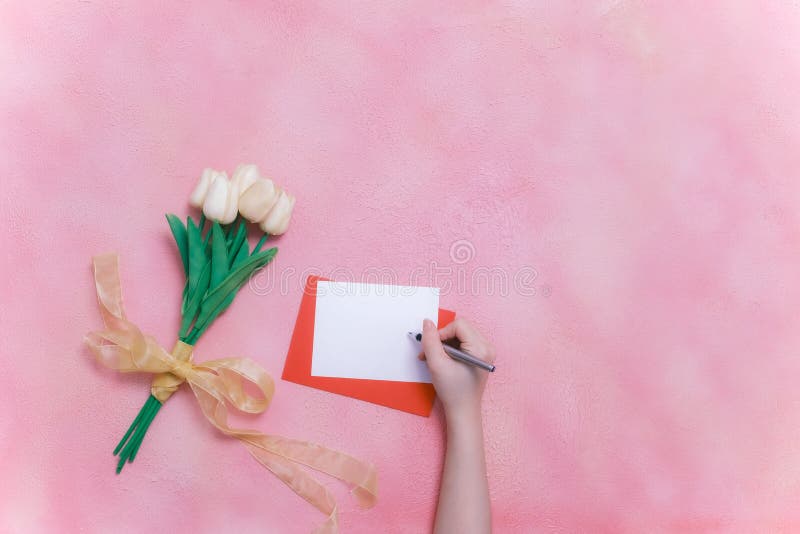 Woman hands holding pen, ready to write. flowers, red envelope with blank paper, pen on pink background. Love concept.