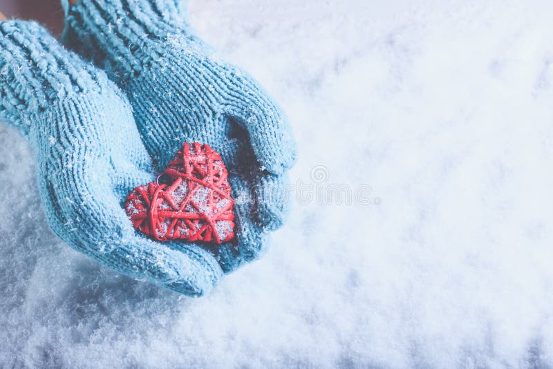 Woman hands in light teal knitted mittens are holding beautiful entwined vintage red heart on snow. Love, St. Valentine concept.
