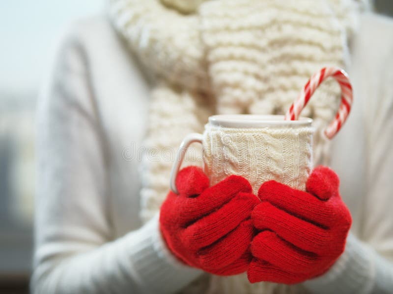 Woman hands in woolen red gloves holding a cozy mug with hot cocoa, tea or coffee and a candy cane. Winter, Christmas time concept