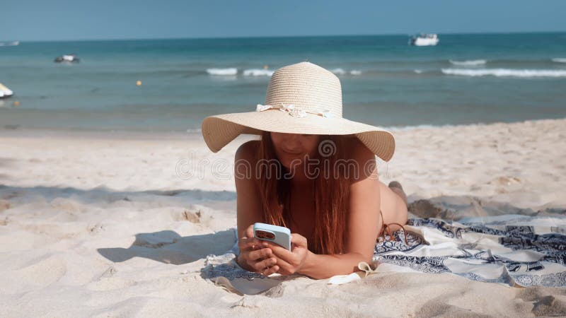 Woman in hat and bikini lying down on sandy beach using mobile phone vacation. Beach vacation ocean's melody in