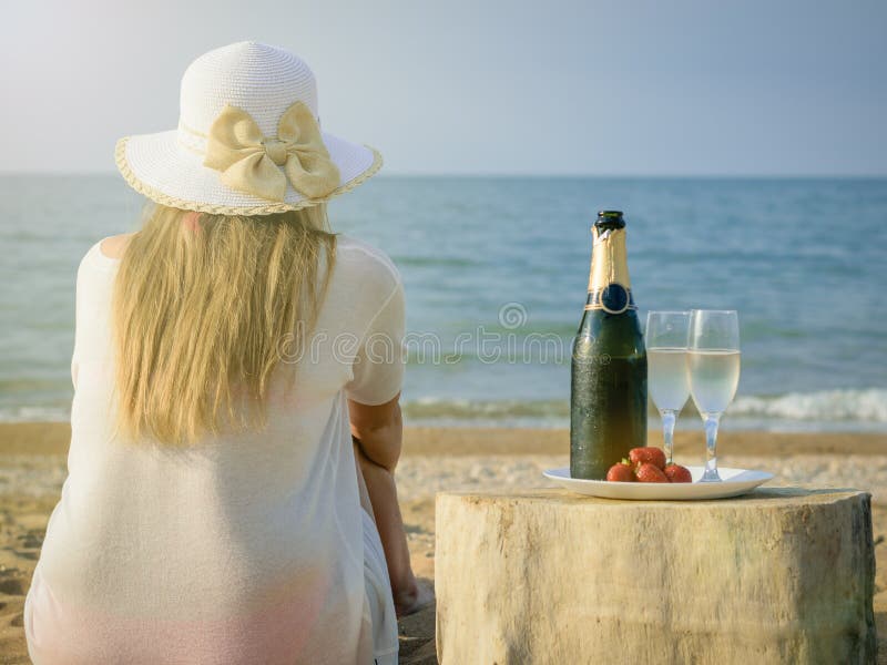 Woman in hat at the sea with an open bottle of champagne and two glasses.