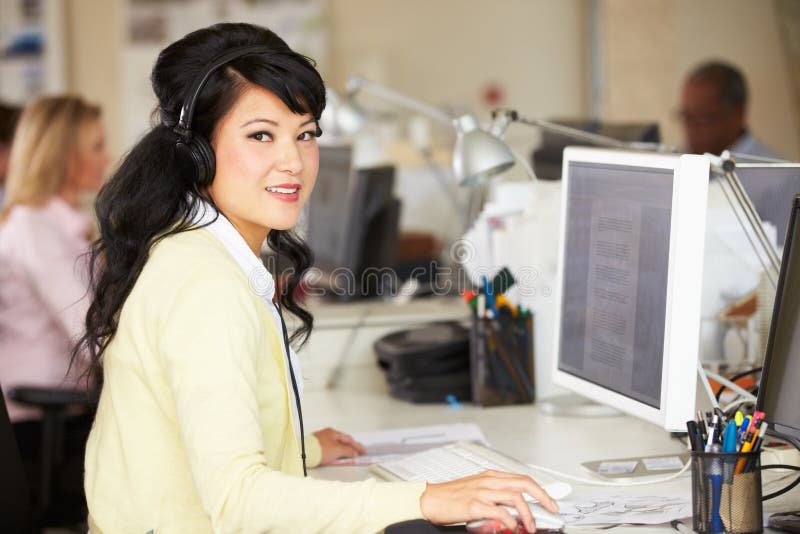 Woman With Headset Working At Desk In Busy Creative Office Smiling