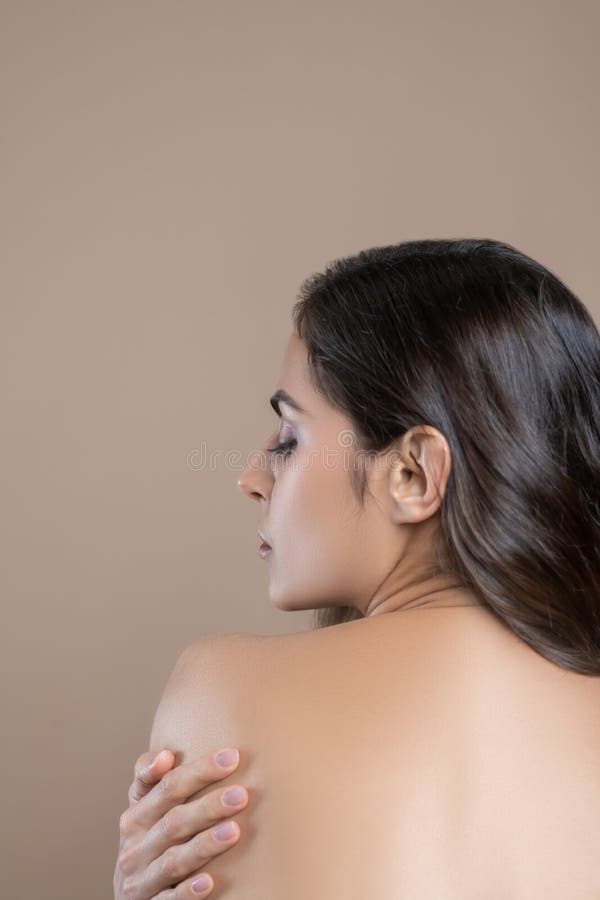 Woman with her back holding her shoulder. Gentle touch. Dark-haired pretty woman with her back holding shoulder with hand and turning face to camera