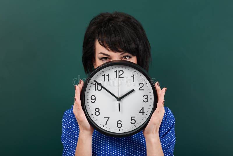 Woman hides her face behind the clock, posing by chalk board, time and education concept, green background, studio shot stock photo Woman hides her face behind the clock, posing by chalk board, time and education concept, green background, studio shot stock photo