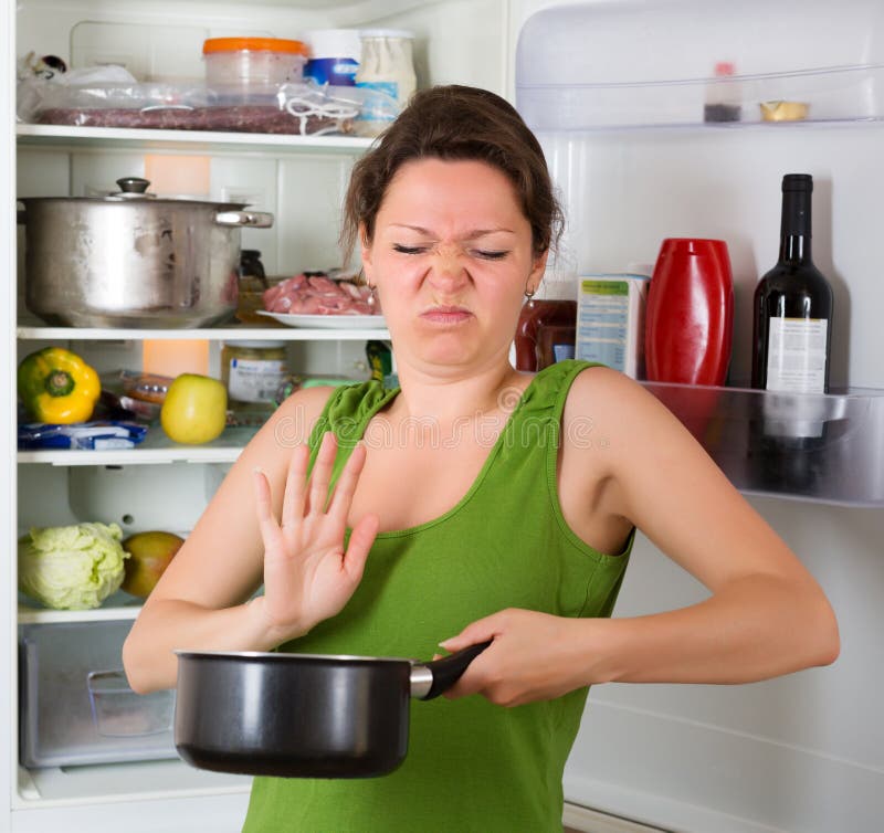 Young woman holding her nose because of bad smell from food near refrigerator at home. Young woman holding her nose because of bad smell from food near refrigerator at home