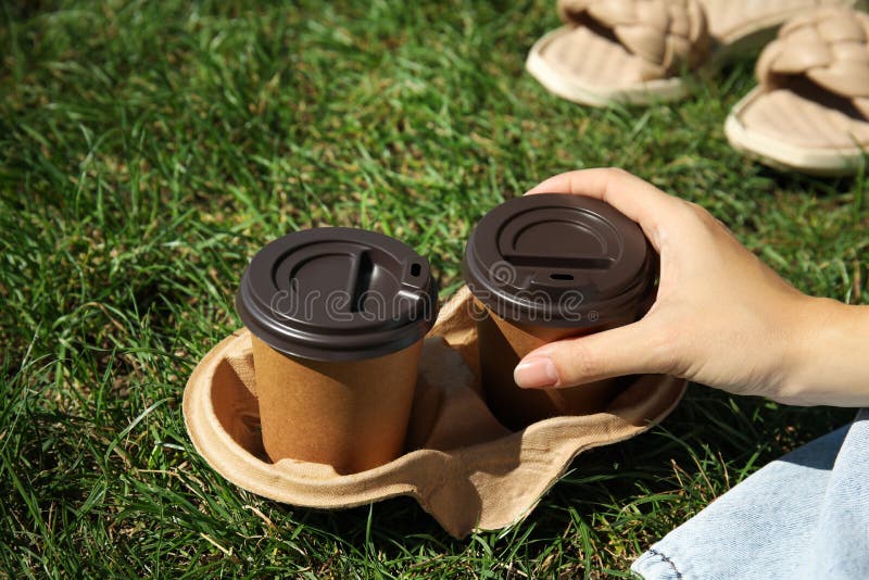 Woman holding takeaway cardboard coffee cups with plastic lids on green grass, closeup stock photo
