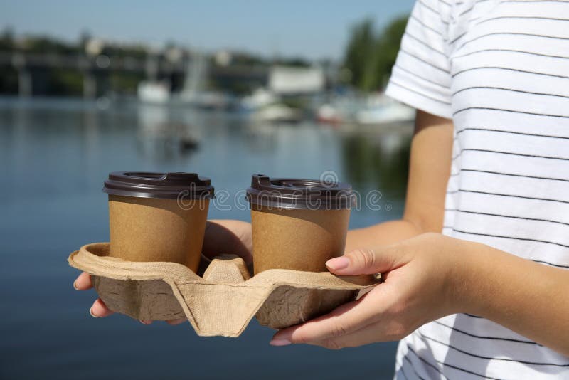 Woman holding takeaway paper coffee cups with plastic lids in cardboard holder near river, closeup stock photography