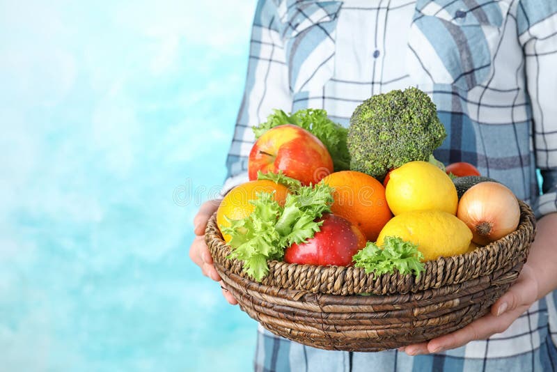 Woman holding wicker bowl with ripe fruits and vegetables on color background, closeup