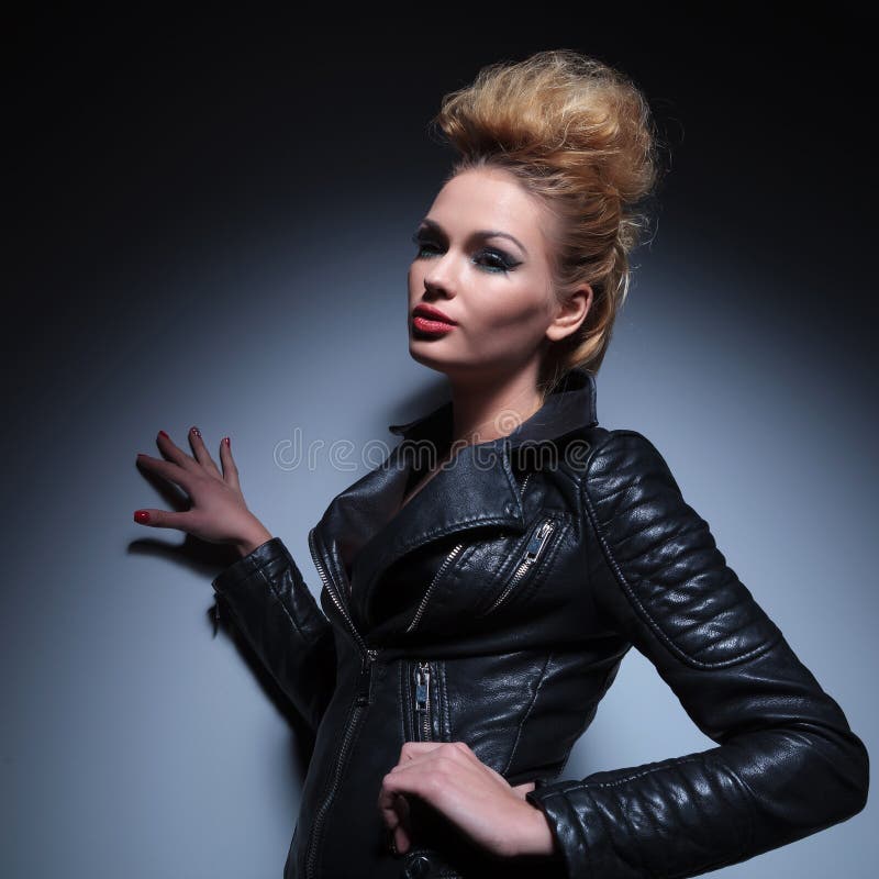 Woman in leather jacket leaning with one hand against a gray stu