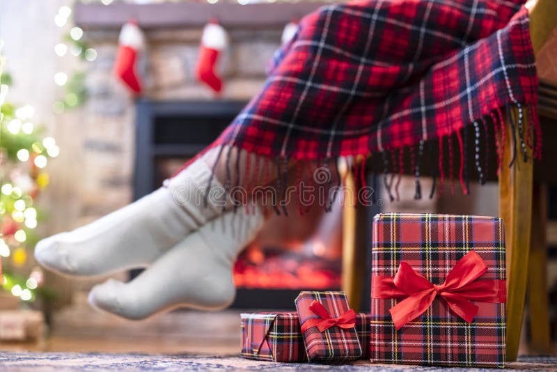 Woman legs in a winter socks covered plaid sitting and relaxation on armchair near fireplace and christmas tree pakking gift boxes