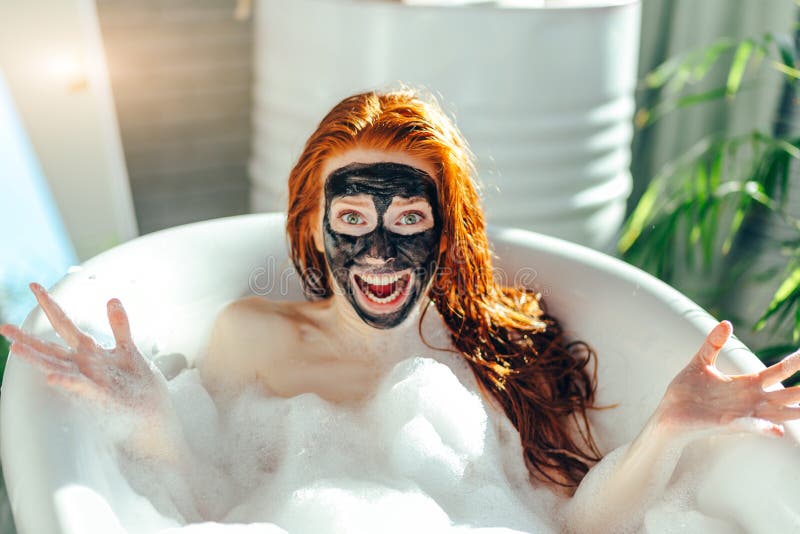 Woman With Long Red Hair Lying In Bathtub With A Clay Mask On Her Face
