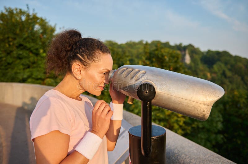 Woman looks through a telescope at the city. Athletic woman in a pink t-shirt is watching the beautiful green nature and the river royalty free stock image