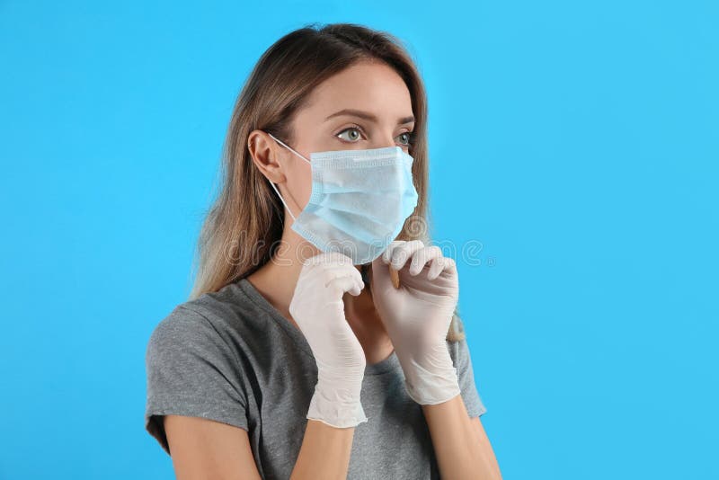 Woman In Medical Gloves Putting On Protective Face Mask Against Light Blue Background Stock