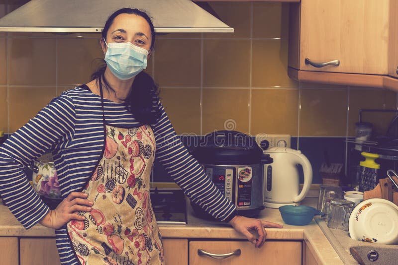Woman in medical mask in the kitchen. Beautiful woman in medical maskin an apron standing in kitchen. toned stock photography Woman in medical mask in the kitchen. Beautiful woman in medical maskin an apron standing in kitchen. toned stock photography