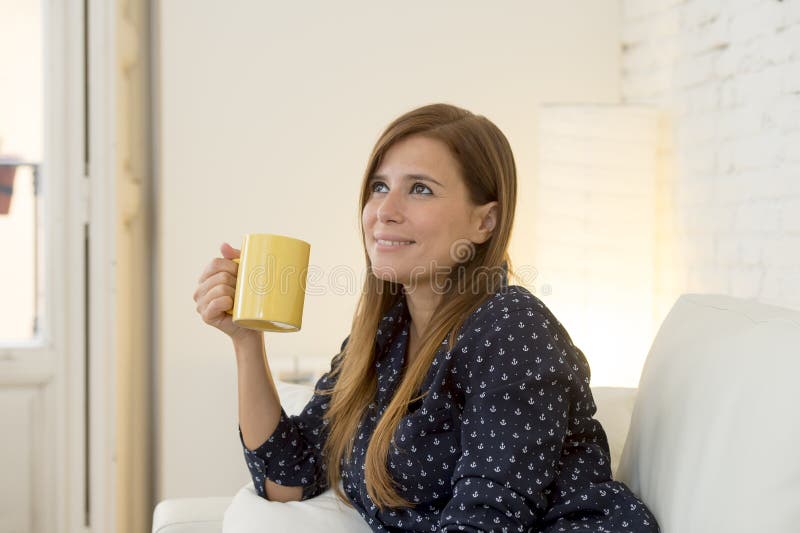 Young attractive woman sitting comfortably at modern apartment living room home couch enjoying a cup of hot tea or coffee smiling happy and relaxed in female domestic lifestyle concept. Young attractive woman sitting comfortably at modern apartment living room home couch enjoying a cup of hot tea or coffee smiling happy and relaxed in female domestic lifestyle concept