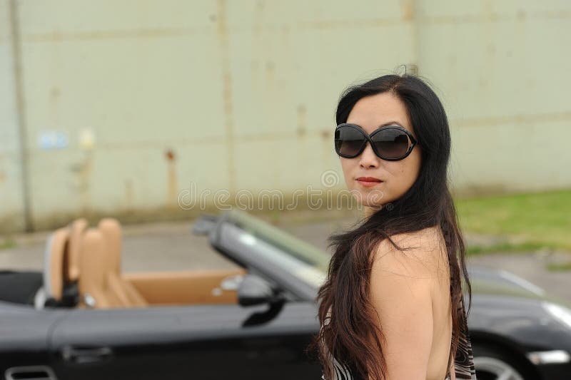 Asian woman with long hair wearing sunglasses next to black sports car. Asian woman with long hair wearing sunglasses next to black sports car.