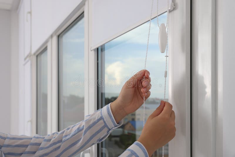 Woman opening white roller blinds on window indoors, closeup