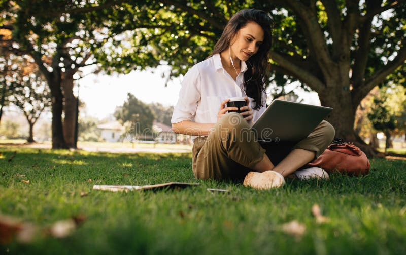 Beautiful woman sitting on grass lawn at park holding a coffee and looking at laptop computer. Young businesswoman in casual clothes sitting at park with coffee using laptop. Female watching video on laptop. Beautiful woman sitting on grass lawn at park holding a coffee and looking at laptop computer. Young businesswoman in casual clothes sitting at park with coffee using laptop. Female watching video on laptop.