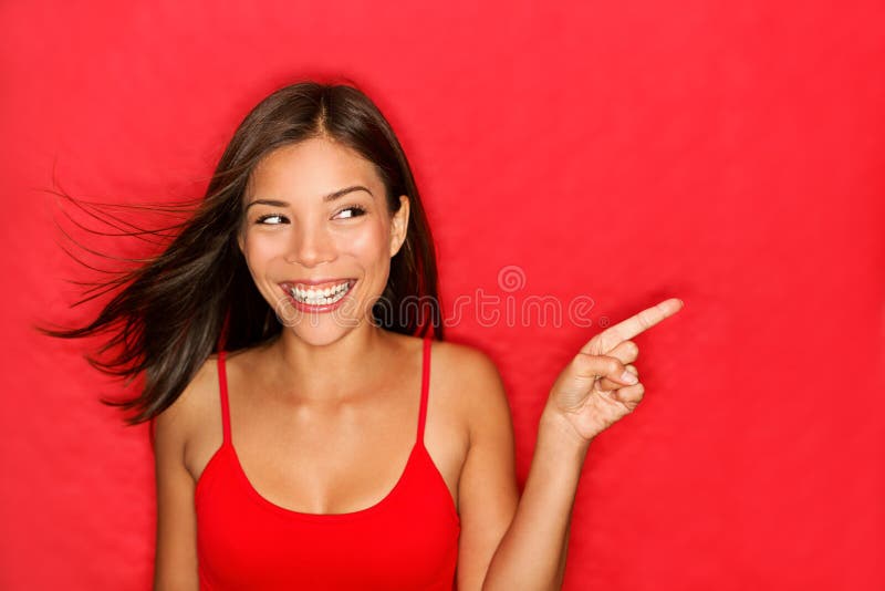 Woman pointing showing on red background looking to the side. Very fresh and energetic beautiful young multiracial Chinese Asian / Caucasian girl smiling happy presenting on red background.