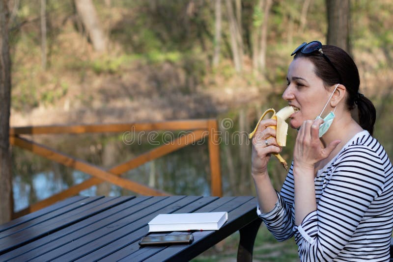 Woman with a protective mask on face, eating a banana..