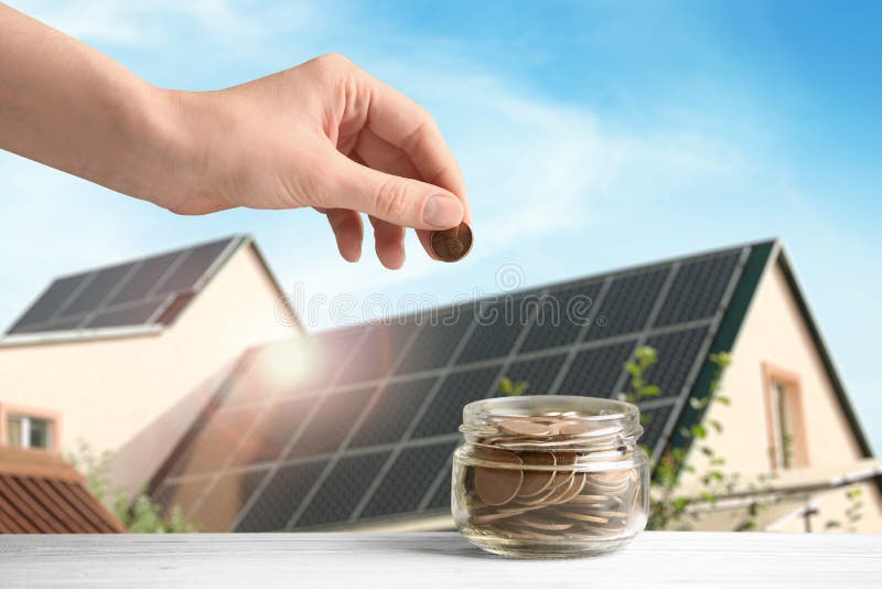 Woman putting coin into jar against house with installed solar panels on roof, closeup. Economic benefits of renewable energy