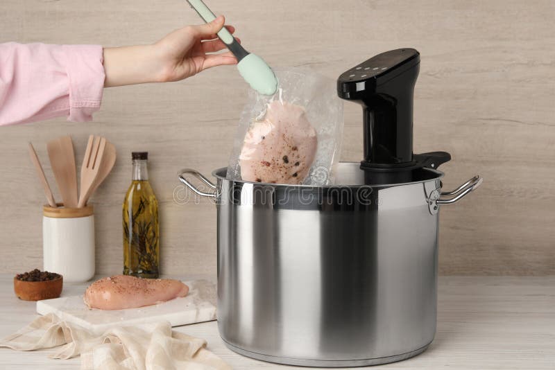 Woman putting vacuum packed meat into pot with sous vide cooker at table, closeup. Thermal immersion circulator