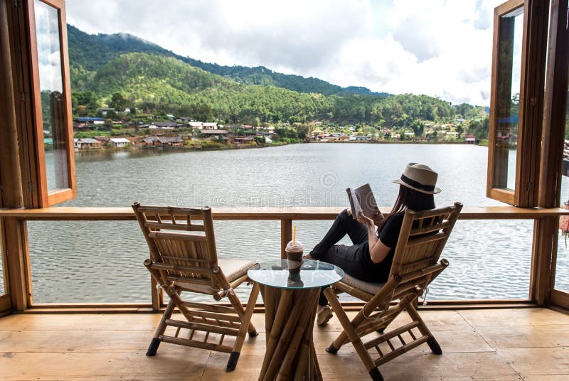 Woman reading a book lying and drink ice coffee on the chair in wood terrace against beautiful view, relax and happy day. Lifestyle Concept