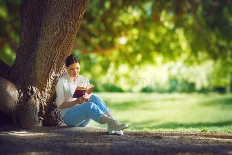 Young woman reading a book under big tree in the park outdoors.