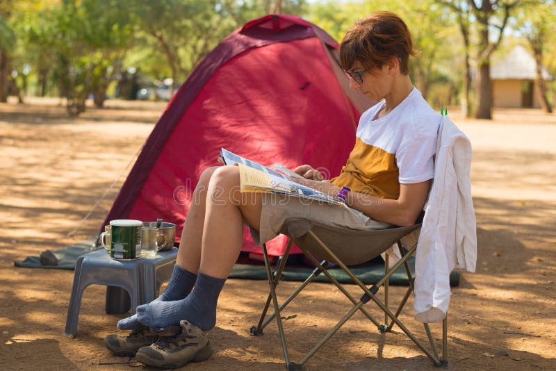 Woman Reading Magazine While Relaxing In Camping Site. Tent, Chairs And Camping Gears. Outdoor Activities In Summer. Adventure Tra