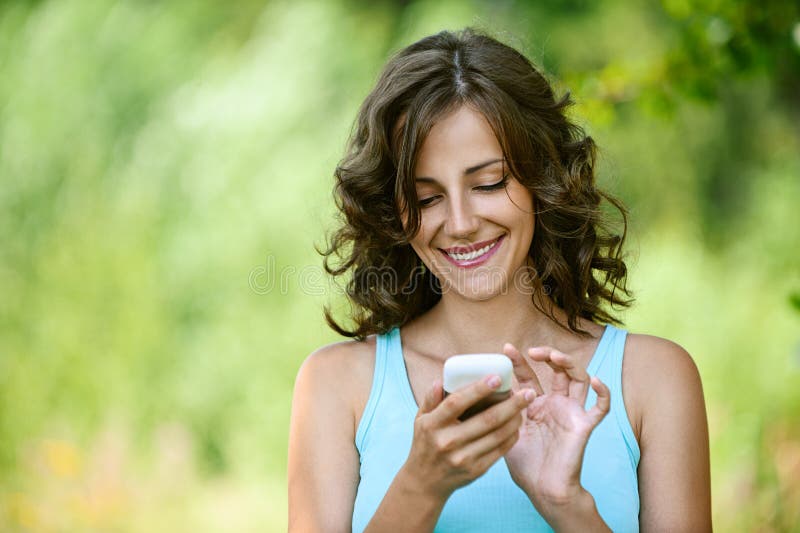 Encantador mujer joven en azul camisas el lee un mensaje sobre el móvil teléfono, contra verde de verano.