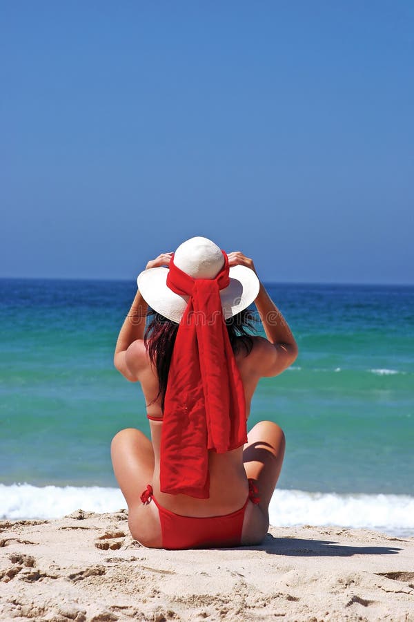 Giovane donna in bikini rosso, seduto sulla spiaggia di sabbia di regolazione cappello e guardando verso il mare.