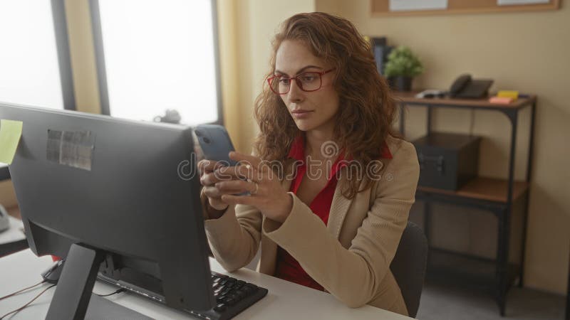 Woman with red glasses in an office using a smartphone at her desk, surrounded by a computer and organizational items, focused on royalty free stock photography