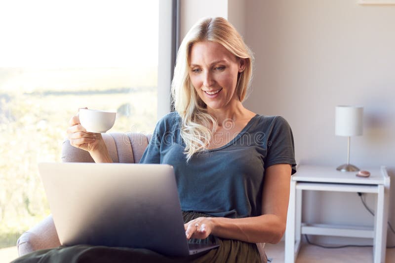 Woman Relaxing In Chair By Window At Home Using Laptop Drinking Coffee stock photography