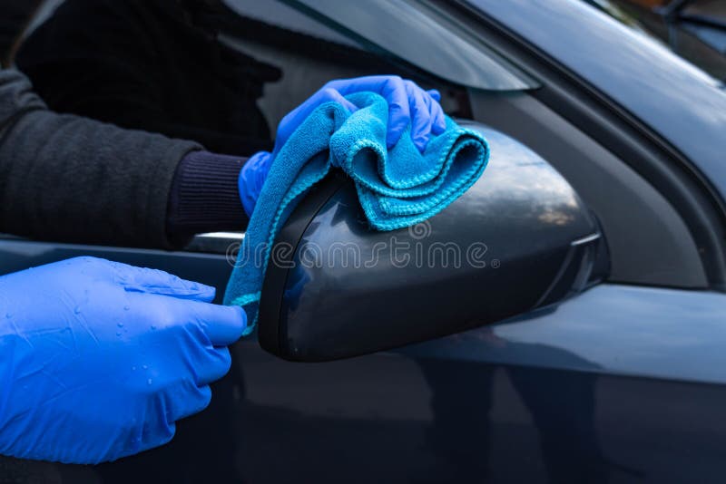 A woman`s hand in a blue glove washes the car mirror with a blue rag. Car care and washing