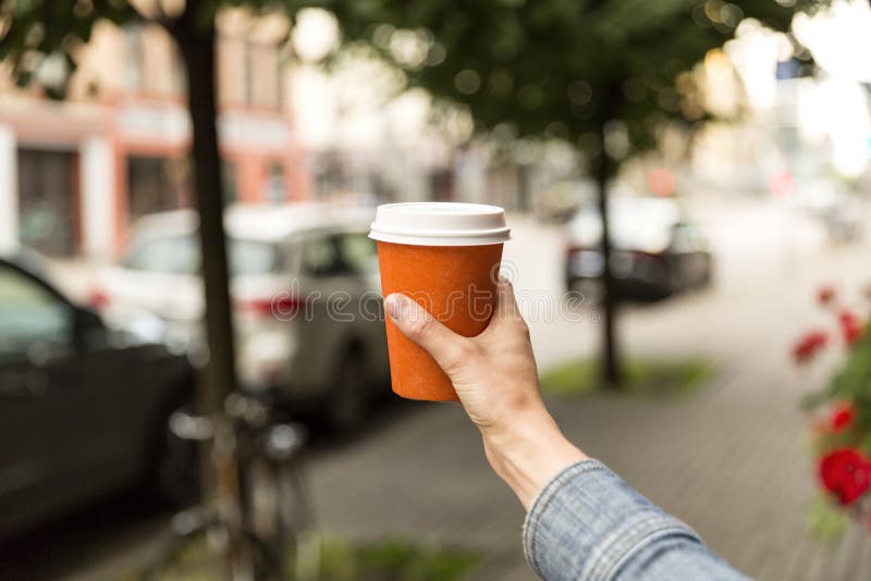 Woman`s hand with coffee in paper cup, city view background