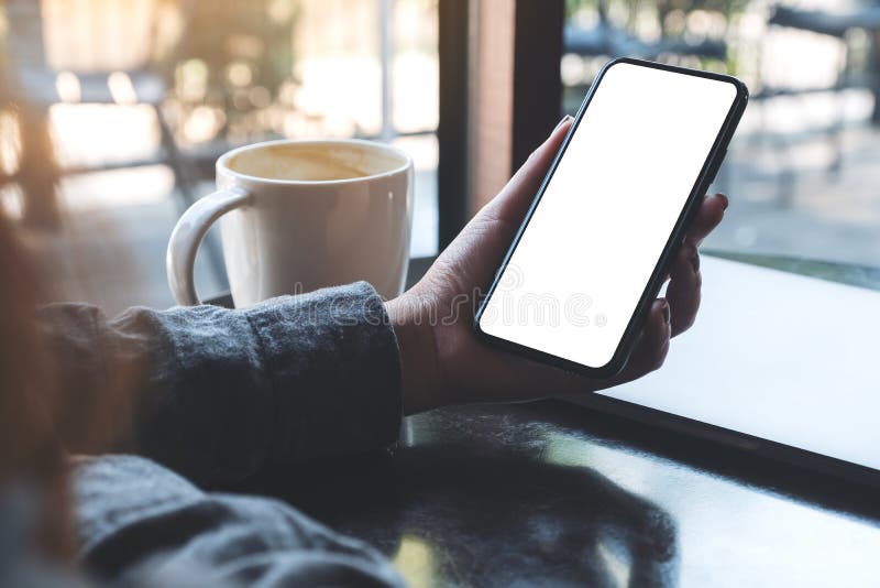 Mockup image of a woman`s hand holding black mobile phone with blank white desktop screen with coffee cup and laptop on table in cafe