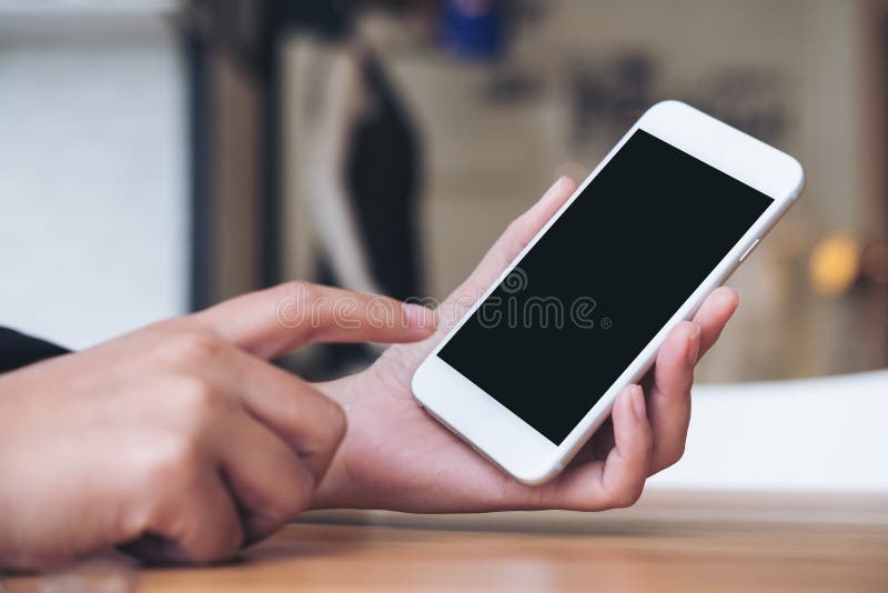 A woman`s hand holding and pointing at white mobile phone with blank black screen in modern cafe