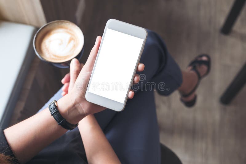 A woman`s hand holding white mobile phone with blank desktop screen while drinking coffee in cafe