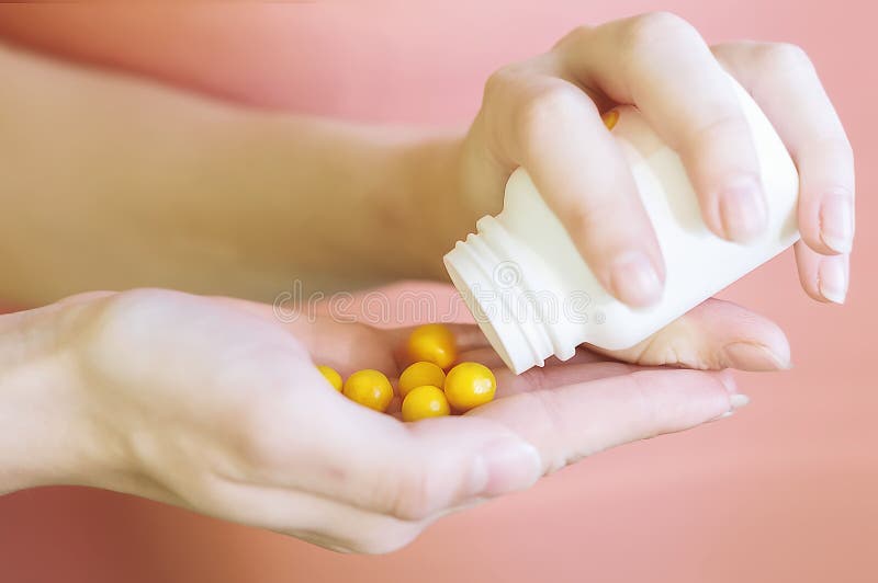 A woman`s hand holds the medicine and pours yellow pills out of the bottle. Hands of a young girl holding a white jar with round vitamins on a pink background. The concept of health, homeopathy.