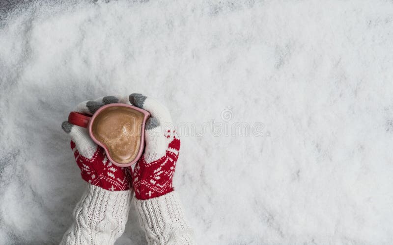 Woman`s hands in Christmas knitted gloves with a cup of coffee in the form of heart on snow. New Year, Christmas and winter conce