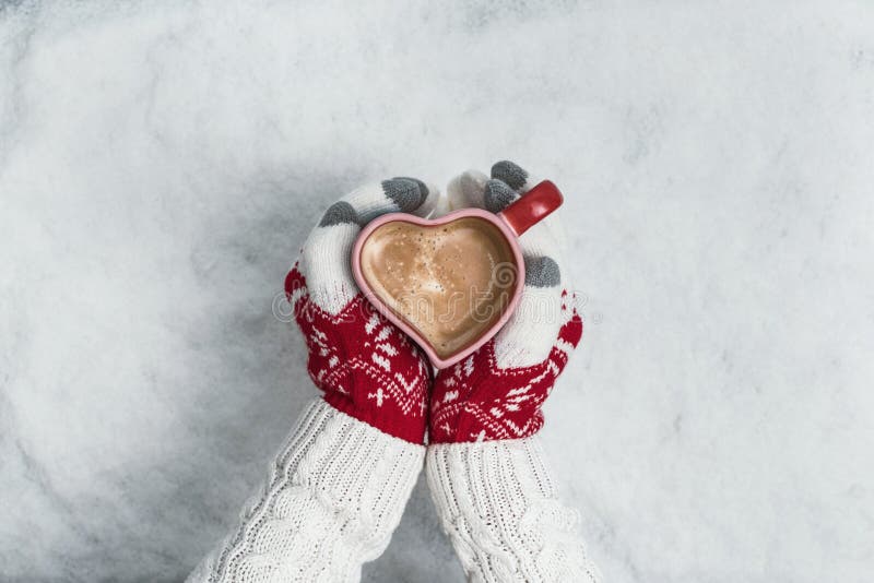 Woman`s hands in Christmas knitted gloves with a cup of coffee in the form of heart on snow. New Year, Christmas and winter conce