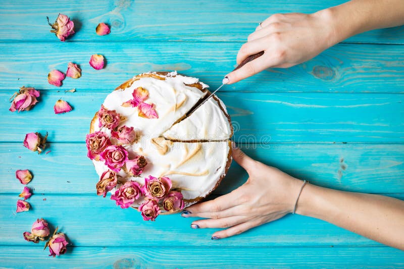 Woman`s hands cut the cake decorated dry rose on blue wood background. Top view.