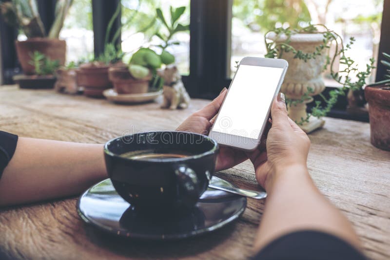 Woman`s hands holding white mobile phone with blank desktop screen and coffee cup on table in cafe