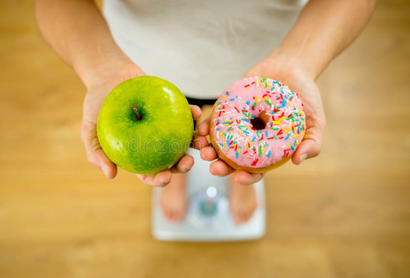 Woman on scale measuring weight holding apple and donuts choosing between healthy or unhealthy food