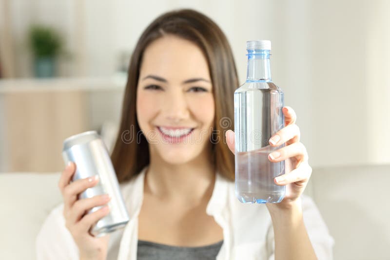 Front view portrait of a happy woman showing a bottle of water and a soda can sitting on a couch in the living room at home. Front view portrait of a happy woman showing a bottle of water and a soda can sitting on a couch in the living room at home