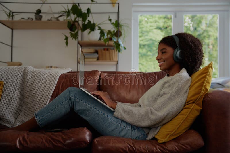 Attractive young woman using a laptop while listening to music on headphones and relaxing on a sofa at home stock images