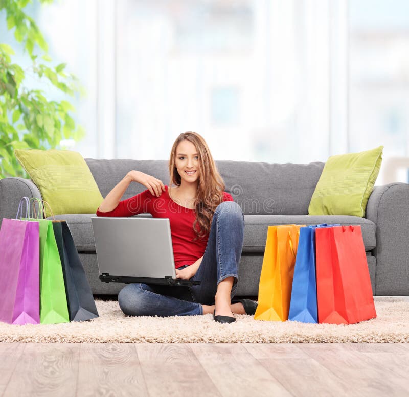 Young woman holding a laptop and sitting in front of a couch with a bunch of shopping bags around her at home shot with tilt and shift lens. Young woman holding a laptop and sitting in front of a couch with a bunch of shopping bags around her at home shot with tilt and shift lens