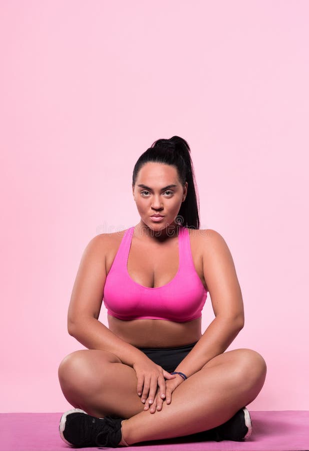 Very serious. Portrait of concentrated young chubby female sitting on floor with crossed legs against pink isolated background. Very serious. Portrait of concentrated young chubby female sitting on floor with crossed legs against pink isolated background.