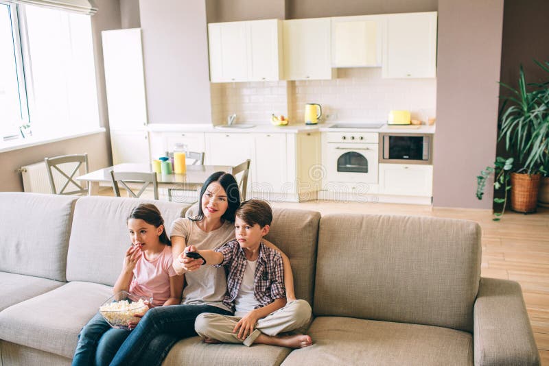 Woman is sitting with her kids and looking straight. Girl eats popcorn. Boy is using remote control from TV. Woman holds royalty free stock photo Woman is sitting with her kids and looking straight. Girl eats popcorn. Boy is using remote control from TV. Woman holds royalty free stock photo